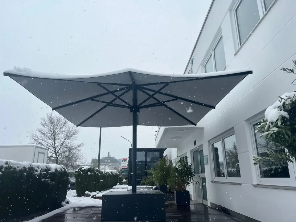 Geöffneter dunkler Großschirm mit Schnee auf dem Dach neben dem Firmengebäude, es schneit