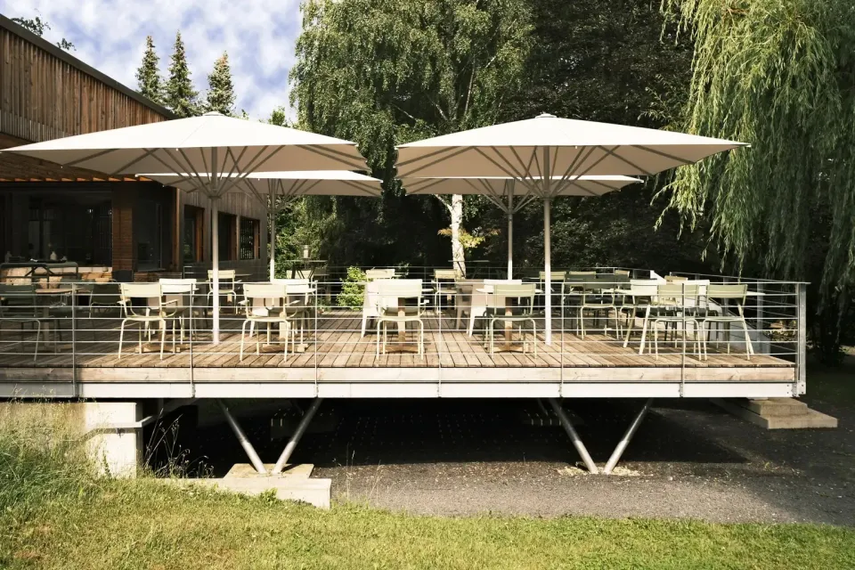 Holzterrasse auf Stützen mit vier weißen Großschirmen über Tischen und Stühlen, dahinter Bäume und ein Holzhaus