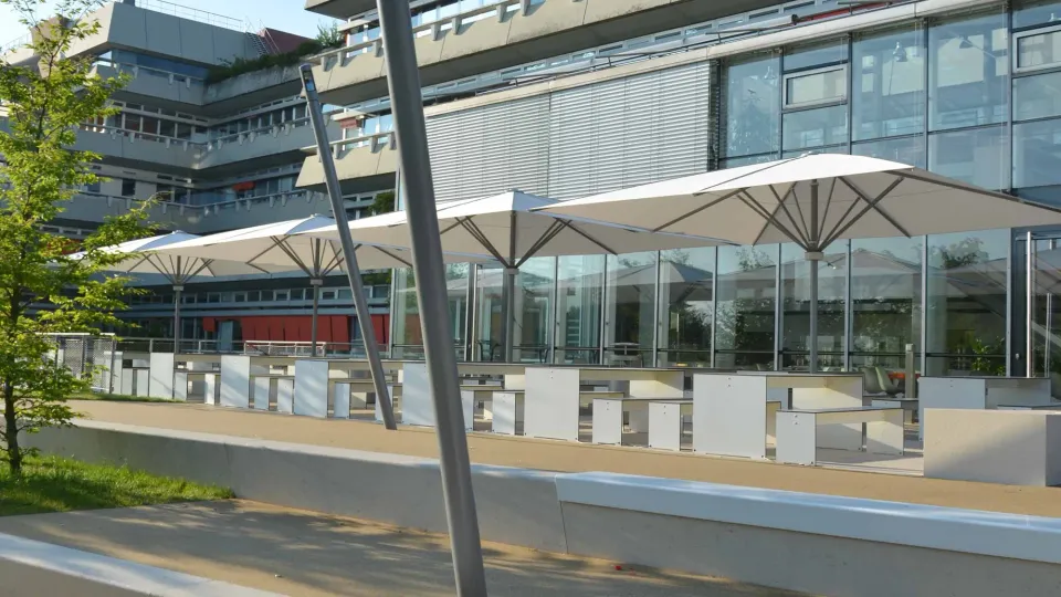 Reihe weißer Großschirme über langen Tischreihen auf einer schmalen Terrasse vor einer Glasfassade