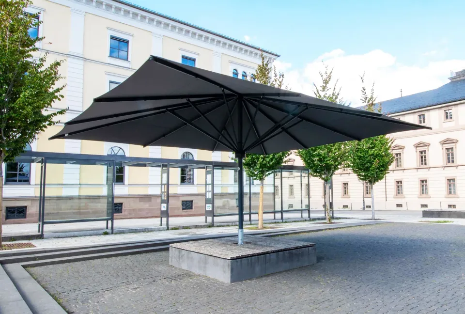 Dunkler quadratischer Großschirm auf einem Steinsockel an einem Stadtplatz mit Wartehäuschen