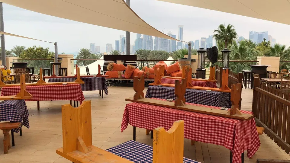 Beiges Segel über einer Restaurantterrasse mit rot-karierten Tischdecken und Holzbänken, im Hintergrund eine Skyline