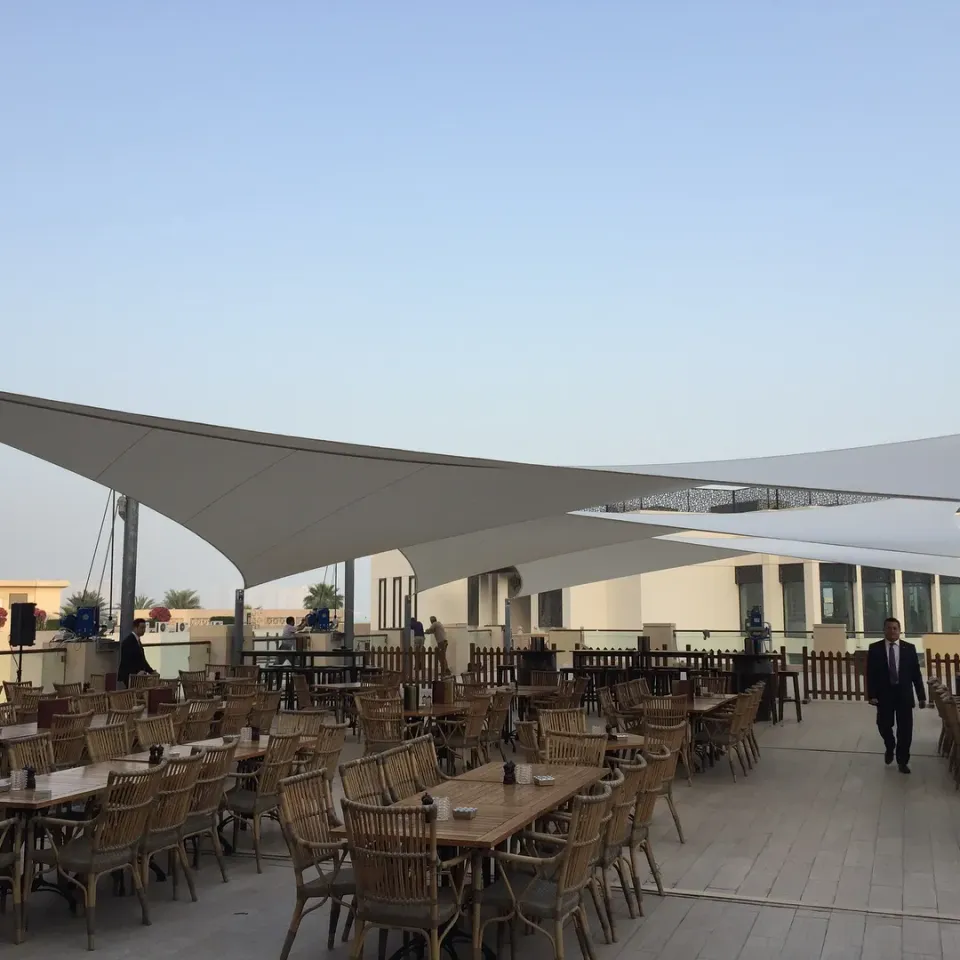 Weiße Segel über einer Terrasse mit Holztischen und Rattanstühlen unter hellem Himmel