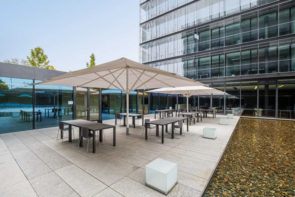 Zwei helle Großschirme über der Terrasse eines Bürogebäudes mit Glasfassade, daneben ein Wasserbecken mit Kieseln