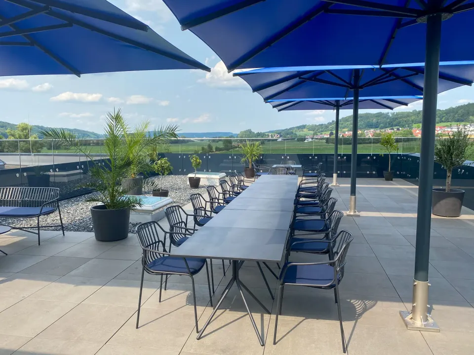 Blaue Großschirme über einer Dachterrasse mit langem Tisch und Stühlen, dahinter weite Landschaft