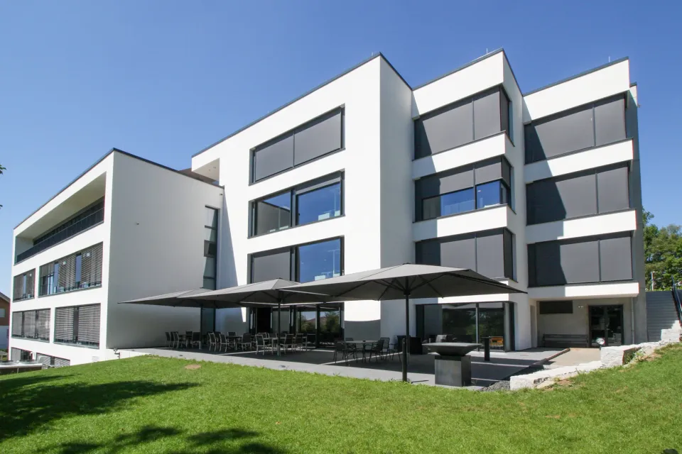 Mehrere dunkle Großschirme über einer Terrasse an einem weißen Bürogebäude, davor eine Rasenfläche