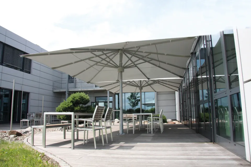 Großer grauer Schirm über einer Terrasse mit Holzdeck vor einem Bürogebäude mit Glasfassade
