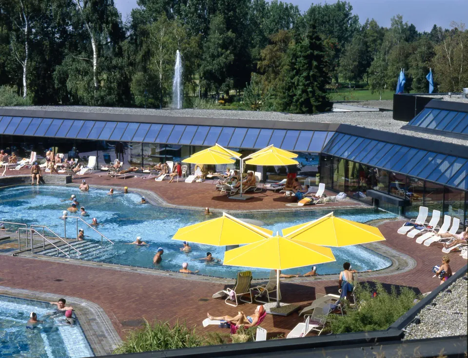 Gelbe Großschirme auf dem Beckenumgang eines Thermalbads, Badegäste im Becken, dahinter Glasfassade und Bäume