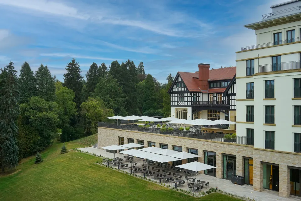 Weiße Großschirme auf mehreren Terrassenebenen vor einem Hotelgebäude mit Park