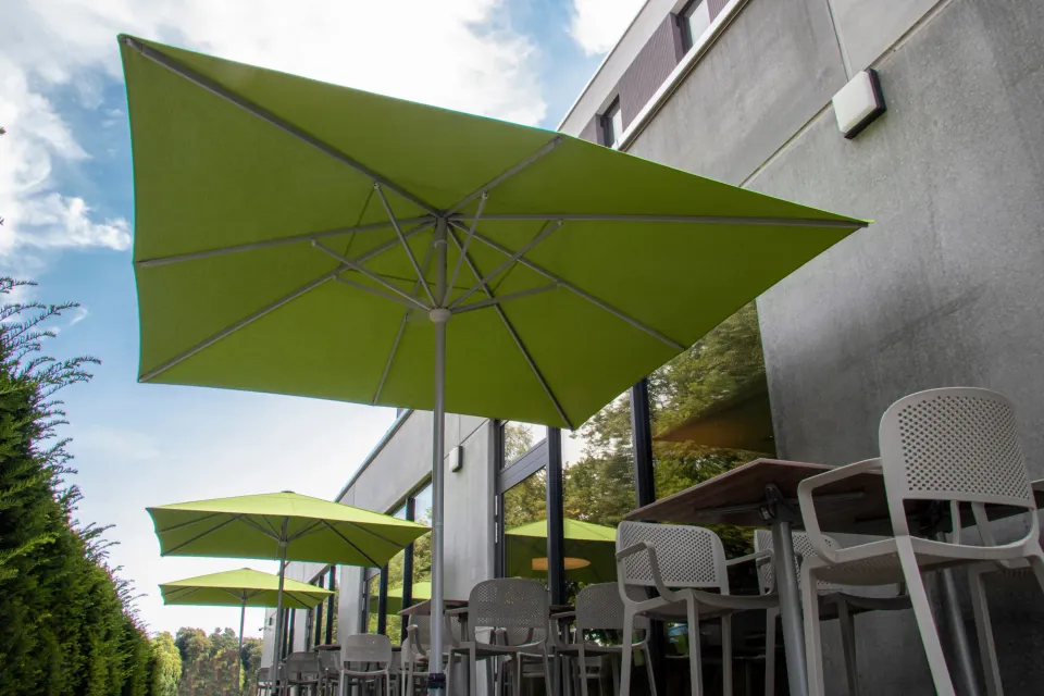 Grüner quadratischer Schirm über einer Terrasse mit weißen Stühlen, dahinter weitere grüne Schirme