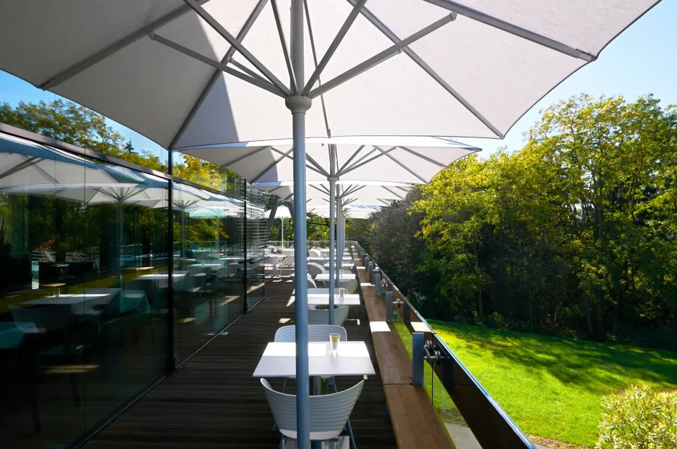 Reihe weißer Schirme über einer Terrasse an einer Glasfront, dahinter Wald und Wiese