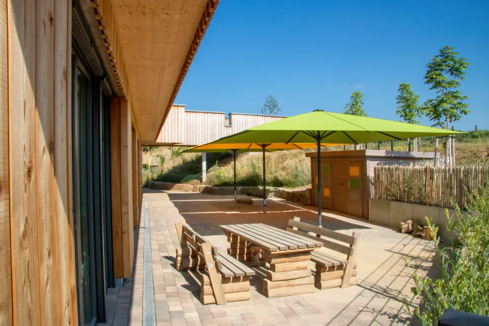 Grüner Großschirm über Holzbänken und Tisch auf der Terrasse eines Kinderhauses