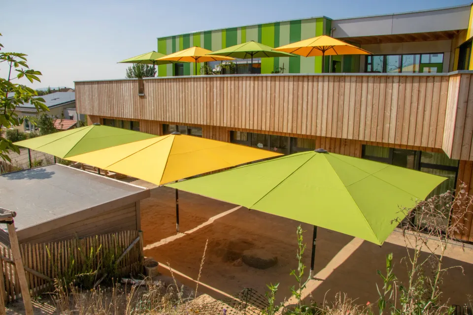 Gelbe und grüne Großschirme über der Holzterrasse eines Kinderhauses, dahinter Sandfläche und farbige Fassade