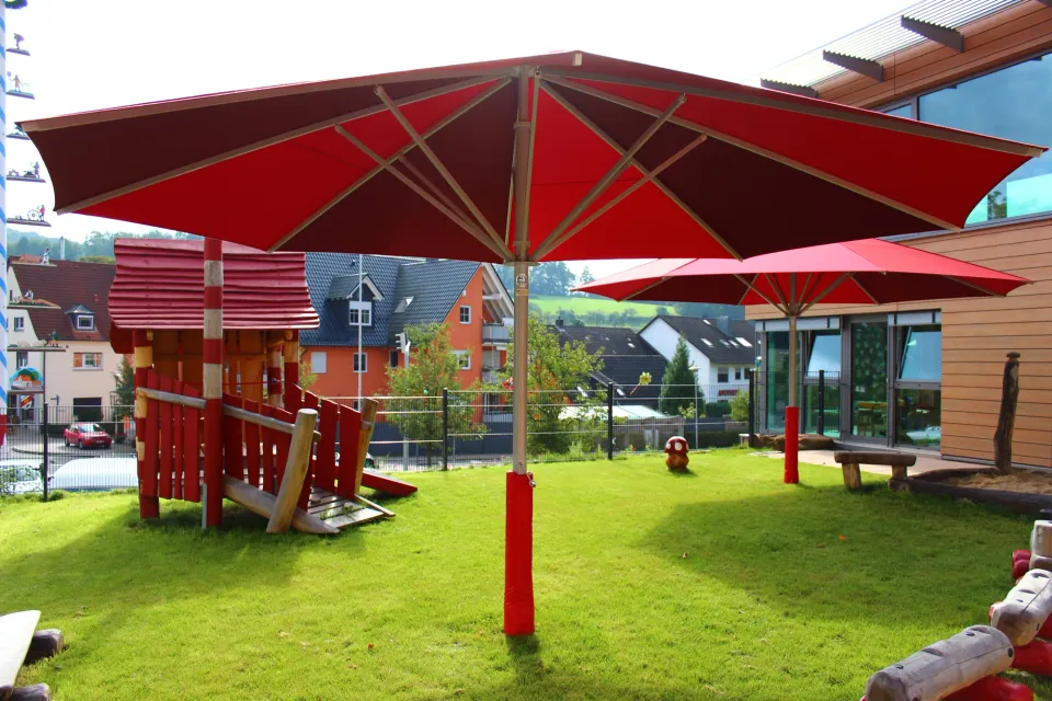 Roter Großschirm mit rotem Prallschutz am Mast neben Spielhaus und Rutsche