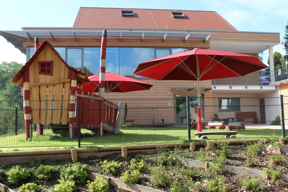 Roter Großschirm mit rotem Prallschutz am Mast neben einem hölzernen Spielhaus im Garten einer Kita