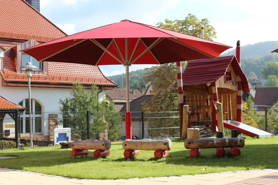 Roter Großschirm neben einem Spielhaus mit Rutsche im Außengelände einer Kita, am Mast ein roter Prallschutz