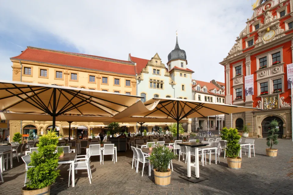 Große cremefarbene Schirme über Tischen und Pflanzkübeln auf einem Marktplatz vor historischen Fassaden und einem Rathaus