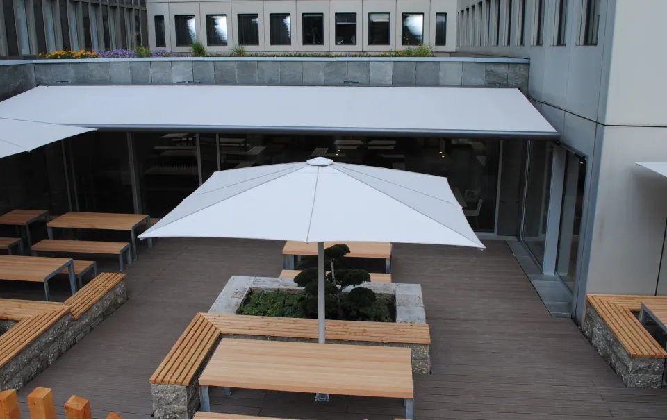 Weißer Schirm über Sitzbänken aus Holz und Gabionen auf der Terrasse eines Firmengebäudes