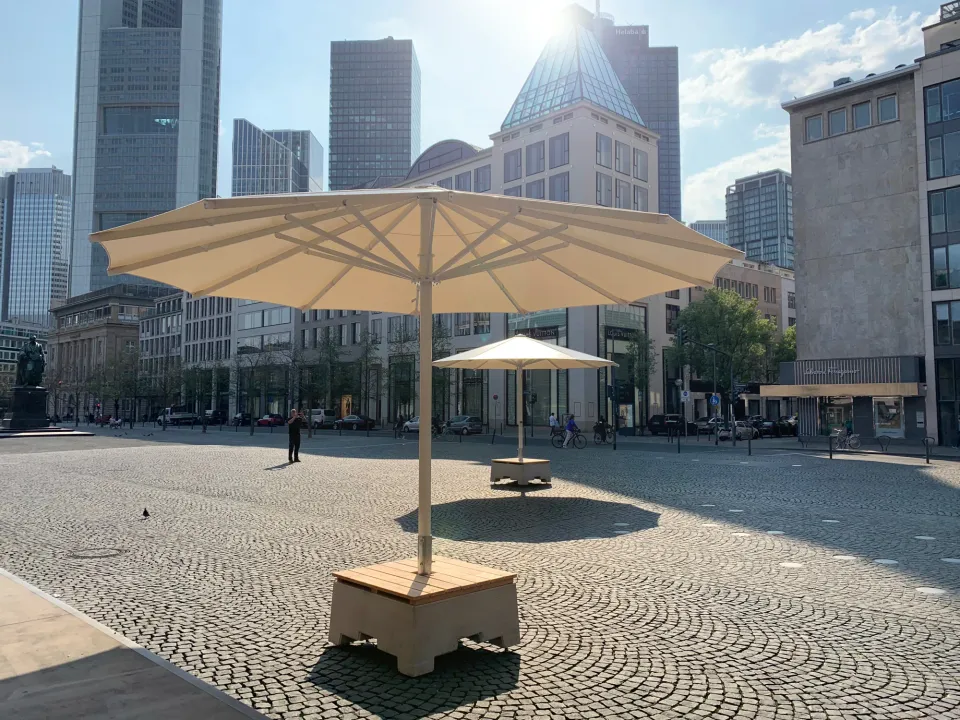Großschirm auf einem Sitzsockel mit Holzauflage auf einem gepflasterten Platz, Hochhäuser im Hintergrund