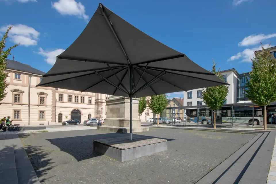 Großer dunkler Schirm über einem Steinsockel an einer Treppe auf einem gepflasterten Platz mit Gebäuden im Hintergrund