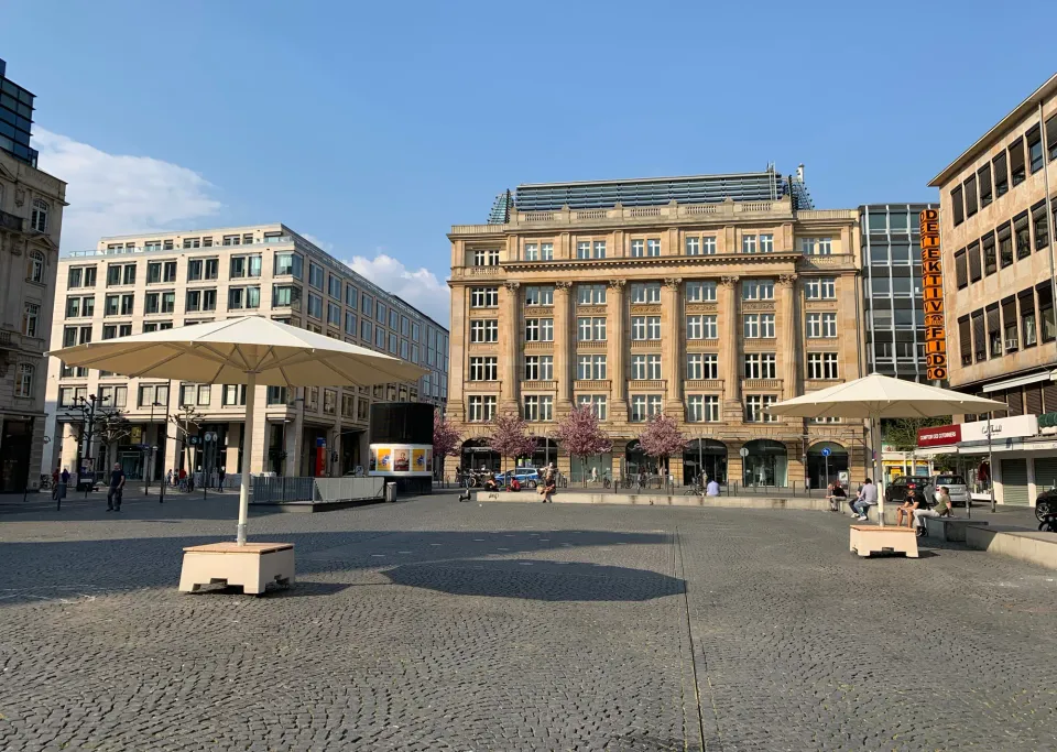 Zwei Großschirme auf Sitzsockeln mit Holzauflage auf einem gepflasterten Stadtplatz, im Hintergrund Geschäftshäuser