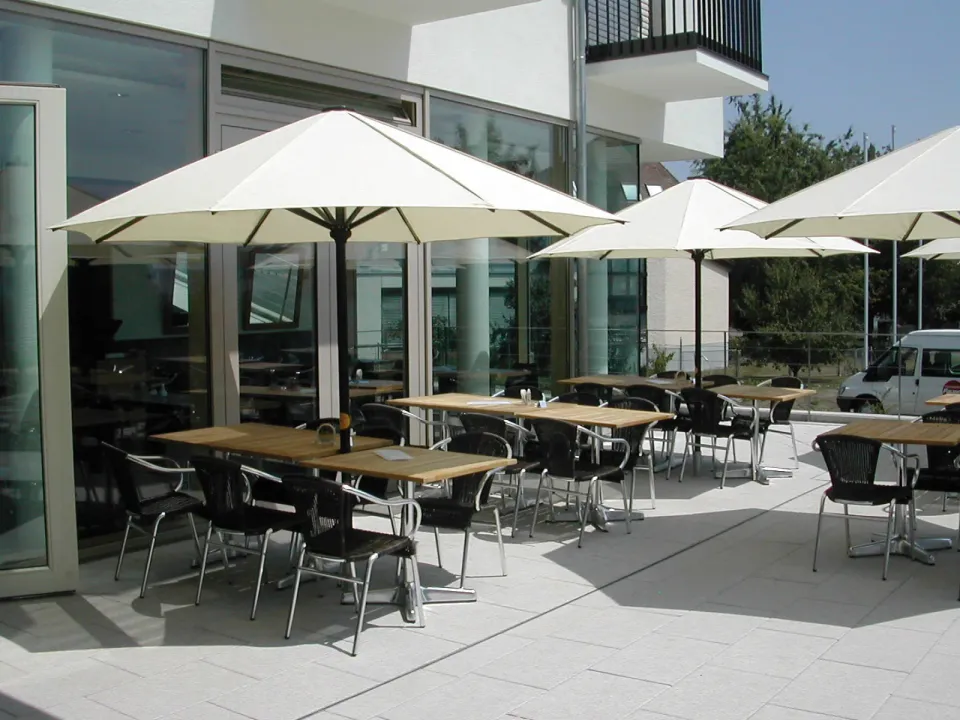 Runde weiße Schirme über Tischen mit Holzplatte auf einer Terrasse vor einer Glasfassade