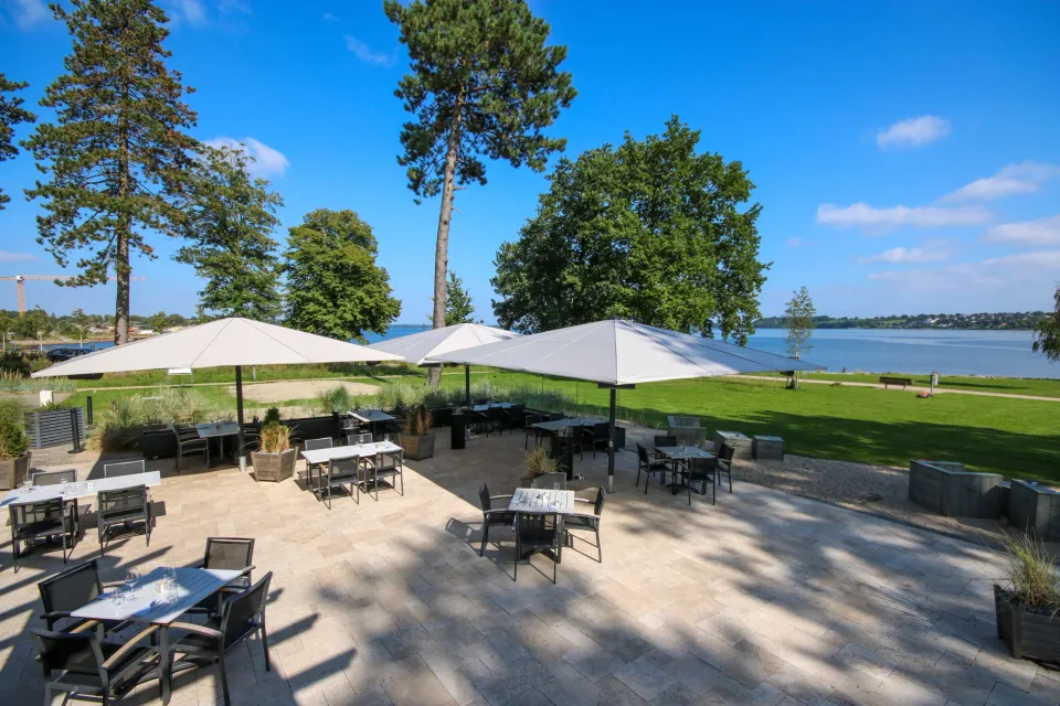 Großschirme über einer gepflasterten Terrasse am See, Tische und Stühle, Kiefern und Wasser im Hintergrund