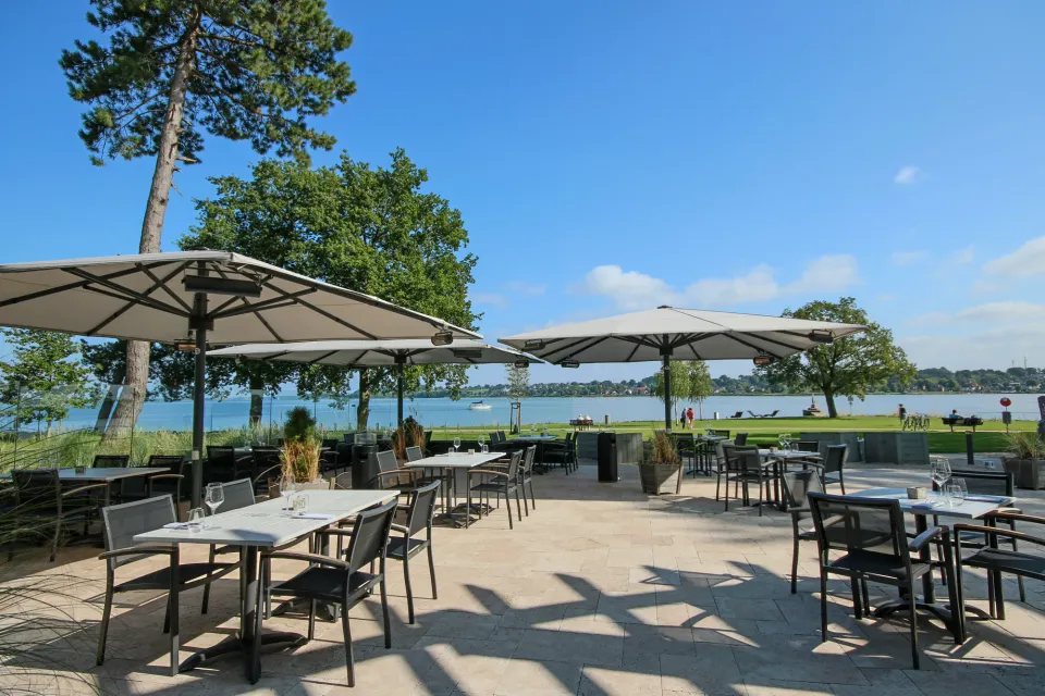 Großschirme mit Heizstrahlern über einer gepflasterten Terrasse am See, Kiefern und Wasser im Hintergrund