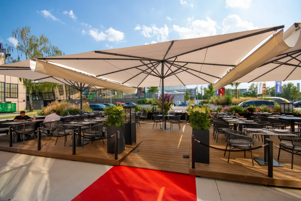 Große cremefarbene Schirme über einer Restaurantterrasse mit Holzdeck, Pflanzkübeln und rotem Läufer