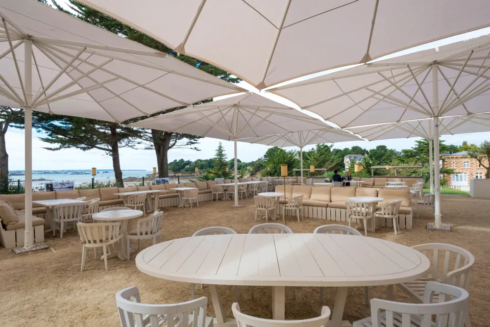 Weiße Großschirme über einer Terrasse mit Sitzbank, Tischen und Stühlen, dahinter Kiefern und Meerblick
