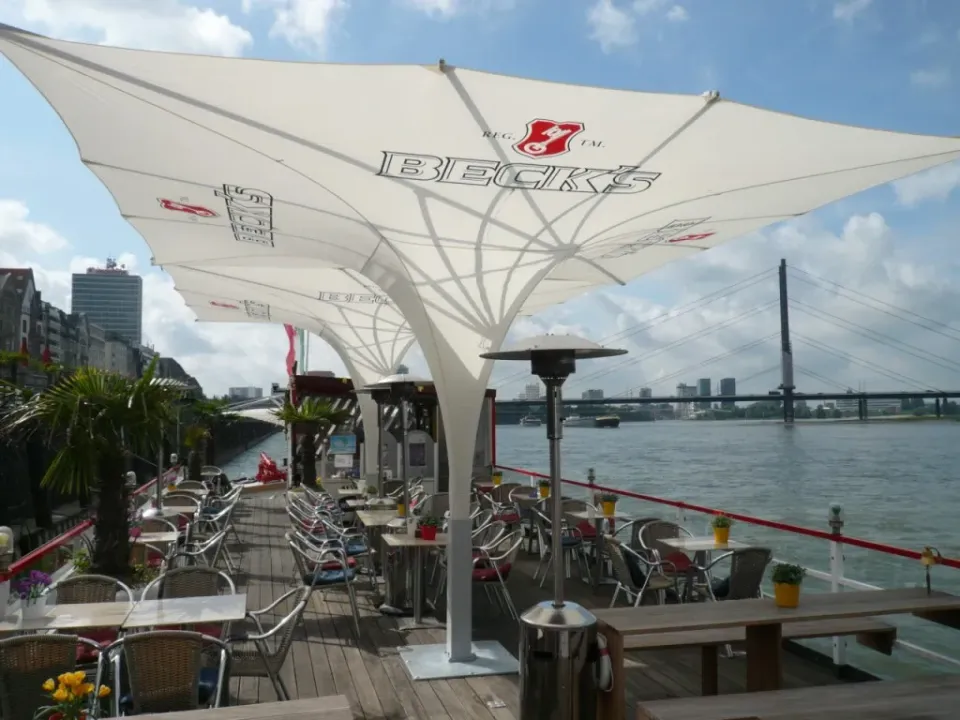 Bedruckter weißer Tulpenschirm über einer möblierten Gastronomieterrasse am Wasser