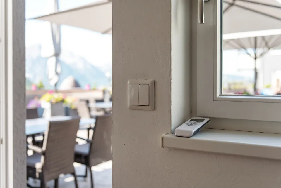 Funk-Fernbedienung auf der Fensterbank neben einem Wandschalter, dahinter eine Terrasse mit Schirmen