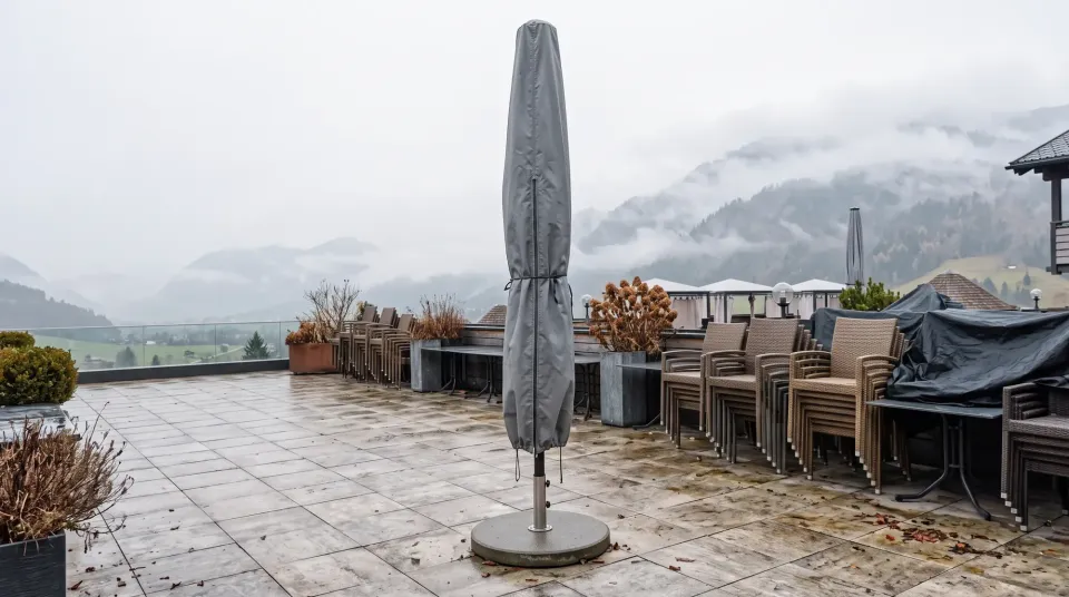 Geschlossener Großschirm in grauer Schutzhülle auf einer Terrasse außerhalb der Saison, Möbel gestapelt